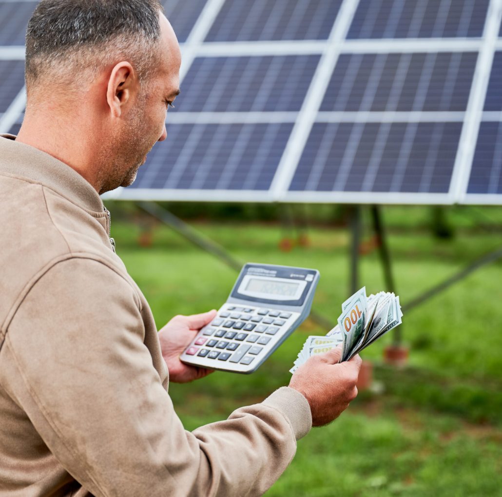 Confident investor counting profit he got from investing in green energy. Glad man holding huge sum received by successfull investment. Back view of man with calculator and money next to PV battery.